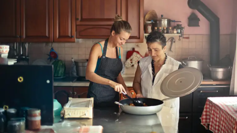 two women cooking together in kitchen putting ingredients in skillet