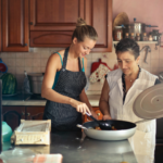 two women cooking together in kitchen putting ingredients in skillet